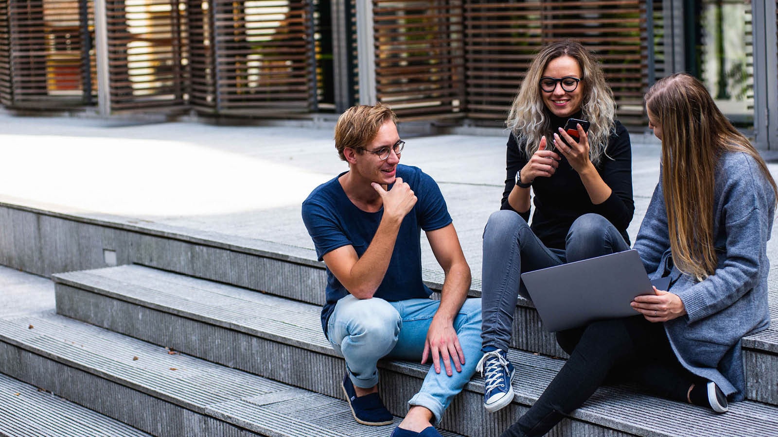 Three young people working outdoors, sitting on steps at the entrance of a building.