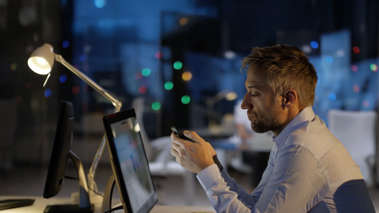 A profile of a man reading from a phone in front of a computer monitor